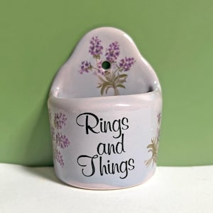 May include: A light purple ceramic wall-mounted holder featuring a painted lavender floral design and the black script text Rings and Things. The piece has a rounded back with a small hole for hanging and a curved front basin for storage.