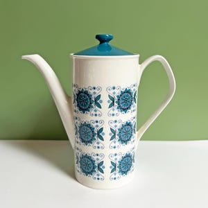 Puede incluir: Una cafetera vintage de cerámica blanca con tapa y asa color turquesa. La cafetera está decorada con un patrón repetido de diseños florales turquesa. El pico es largo y delgado.