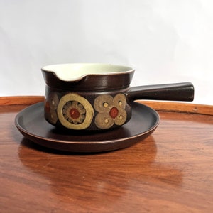 May include: A brown ceramic gravy boat with a white interior and a floral design. The boat has a handle and is sitting on a matching brown saucer.