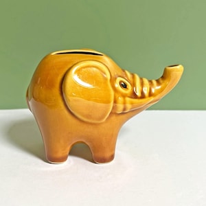 Puede incluir: Una alcancía con forma de elefante de cerámica en un tono marrón dorado cálido. La alcancía tiene una ranura en la parte superior para insertar monedas y una trompa, orejas y patas detalladas. El elefante tiene un acabado brillante y un ojo pequeño y oscuro.