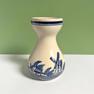 May include: A small, white ceramic vase with a blue and white windmill design. The vase has a rounded body and a narrow neck.