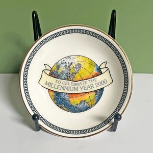 Op de afbeelding: Een klein, rond, wit keramisch bord met een gouden rand en een zwarte binaire code rand. Het bord toont een kleurrijke wereldbol afbeelding met een banner met de tekst "TO CELEBRATE THE MILLENNIUM YEAR 2000".