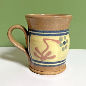 May include: A handmade ceramic mug with a tan exterior and a yellow interior. The mug features blue stripes, a pink floral design, and a blue and green floral design. The handle is tan.