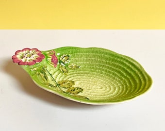 May include: A green ceramic dish with a raised floral design. The dish has a scalloped edge and a textured surface. The flower is pink and has a green stem and leaves.