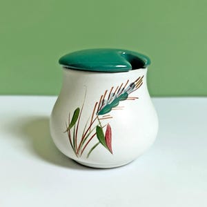 May include: A white ceramic sugar bowl with a green lid and a painted wheat design. The bowl has a round shape and a small spout on the lid.