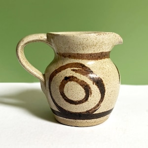 May include: A small ceramic jug with a speckled cream-coloured glaze. The jug features a dark brown hand-painted spiral design on the front, a horizontal brown band near the rim, and a matching brown band around the base with a curved handle.