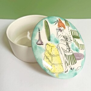 May include: A white ceramic trinket box with an oval lid. The lid features a hand-painted illustration of a woman in a dressing room, with a yellow dress and green and blue accents. The box is open.