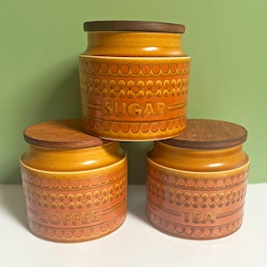 May include: Three cylindrical ceramic storage canisters with dark brown wooden lids. The canisters are glazed in shades of orange and brown, featuring a repeating embossed floral and circular pattern. The words SUGAR, COFFEE, and TEA are embossed.