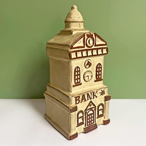 May include: A vintage ceramic bank shaped like a building, with a cream-colored base and brown accents. The word "BANK" is prominently displayed, along with clock details and architectural features. The bank has a domed top and a small door.