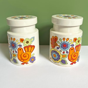 May include: Two white ceramic canisters with matching lids, each featuring a colourful graphic print of an orange bird surrounded by stylised flowers in shades of purple, yellow, orange, and blue. The lids also feature a circular floral pattern.