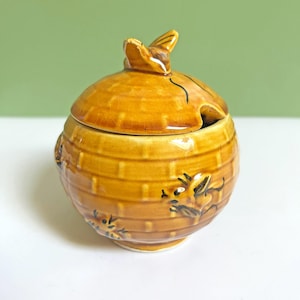 Può includere: Un vasetto per il miele in ceramica di colore giallo-marrone caldo, con coperchio e manico a forma di ape. Il vasetto presenta un motivo a nido d'ape strutturato e dettagli di api dipinte. Il coperchio ha una piccola apertura per un cucchiaio.