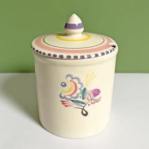 May include: Cream-colored ceramic jar with a lid, featuring a floral design in blue, pink, yellow, and purple. The lid has a decorative rim and a finial with purple stripes. A small spoon rest is on the lid.