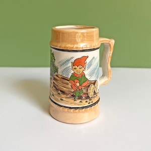 May include: A ceramic beer stein with a brown glaze and a whimsical illustration of a gnome sitting on a log in a forest setting. The gnome is wearing a green and red outfit and has a red cap.