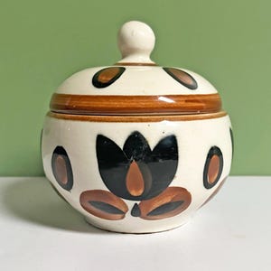 May include: A cream-coloured ceramic jar with a lid. The jar is decorated with brown and black floral patterns. The lid has a small, round knob. The jar is likely used for storing sugar or other small items.