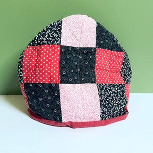 May include: A patchwork tea cozy with a rounded shape. The cozy is made of squares in red with white polka dots, black with white floral patterns, and pink with small floral patterns. A red border trims the bottom.