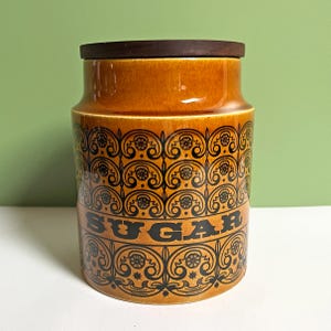 May include: A cylindrical ceramic sugar canister featuring a glossy amber-orange glaze and a dark brown wooden lid. The exterior is decorated with a repeating black scrollwork pattern and the word SUGAR printed in bold black capital letters.