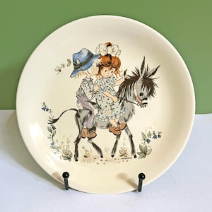 May include: A round white ceramic plate featuring a colorful illustration of a child in a blue hat and a child with a white bow riding a grey donkey. The scene includes small blue flowers and green foliage at the base, with a signature near the donkey.