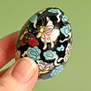 May include: A black, egg-shaped decorative object with colourful butterfly and cloud designs. The butterflies have pink, yellow, and white wings. The clouds are light blue. The object is held in a person's hand.