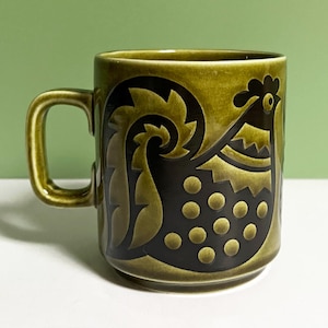 May include: Olive green ceramic mug with a black rooster design. The rooster has gold accents and circular patterns. The mug has a curved handle and a glossy finish. Ideal for tea or coffee, it measures approximately 10 cm in height.