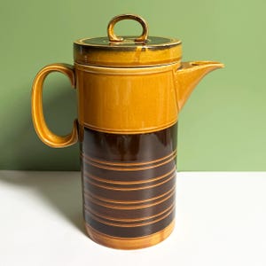 May include: A vintage ceramic coffee pot with a lid. The pot is primarily brown and yellow with horizontal stripes on the lower portion. It has a handle and a spout. The lid has a gold-colored handle.