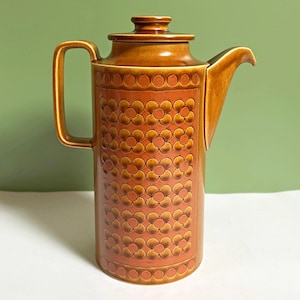 Könnte beinhalten: Eine hohe, zylindrische Kaffeekanne aus Keramik in Braun- und Rostorangetönen. Der Körper zeigt ein sich wiederholendes, geprägtes Blumenmuster und einen glatten Henkel. Sie umfasst einen passenden Deckel mit kleinem Knauf, einen breiten Ausguss und eine glänzende Oberfläche.