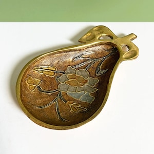 Op de afbeelding: Een decoratieve messing schaal in de vorm van een peer, met een bloemmotief. De schaal heeft een bruin interieur met een bloem in grijstinten, goud en bruin. De rand en de steel zijn goudkleurig.