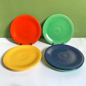 May include: A set of four round ceramic plates featuring concentric ring patterns along the rims. The collection includes one bright orange plate, one light green plate, one yellow plate, and one dark blue plate, all displayed in a two-by-two arrangement.