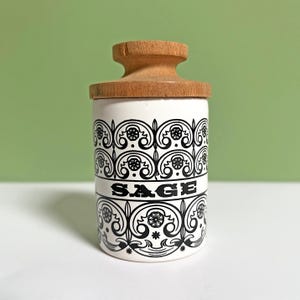 Peut inclure: Un pot en céramique blanc avec un couvercle en bois, orné de motifs noirs et du mot "SAGE". Ce pot est conçu pour la cuisine, probablement pour conserver des herbes ou des épices.