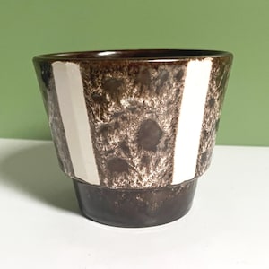 Pode incluir: Um vaso de cerâmica castanho com superfície texturizada e duas faixas brancas verticais. O vaso tem uma base castanha escura e um topo ligeiramente alargado, ideal para pequenas plantas ou suculentas. Mede cerca de 12 cm de altura.