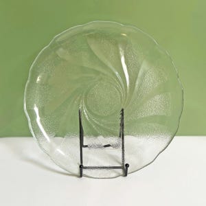 May include: A clear glass plate with a textured surface and a spiral design. The plate has a scalloped edge and is displayed on a black metal stand. The background is a solid green colour.