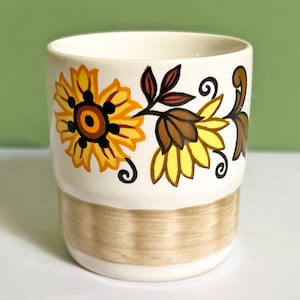 May include: A white ceramic mug with a floral design. The mug features a yellow and brown sunflower pattern with black accents. The base of the mug has a tan band. The mug is cylindrical and appears to be hand-painted.