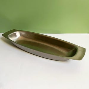 Könnte beinhalten: Eine lange, schmale, oval geformte Servierschale aus Metall mit einer glatten, polierten, silberfarbenen Oberfläche. Das Gefäß verfügt über ein flaches, längliches Becken mit flachen, verlängerten Griffen an beiden Enden für ein elegantes Design.