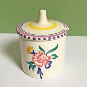 May include: Cream ceramic jar with lid, decorated with a floral design. The lid has a yellow and pink striped pattern and a small handle. The jar is adorned with a pink rose and green leaves. A decorative piece for the home.
