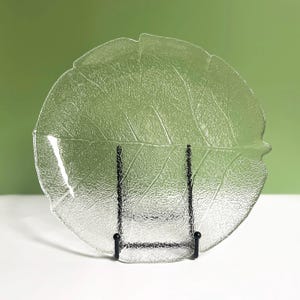 May include: Clear glass leaf-shaped serving dish with textured detailing. The dish is displayed on a black metal stand, showcasing its intricate leaf vein design. The dish is a decorative piece for serving food.