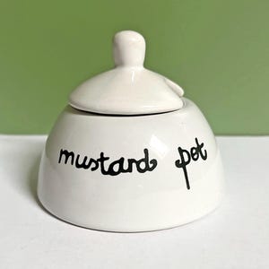 May include: A white ceramic mustard pot with a lid and a handle. The words "mustard pot" are written in black script on the front of the pot. The background is a solid green color.