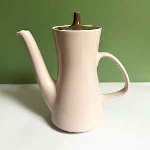 Könnte beinhalten: Eine hellcremefarbene Kaffeekanne aus Keramik mit einem hohen, schlanken Körper, einem geschwungenen Henkel und einem langen, spitz zulaufenden Ausguss. Der flache Deckel hat einen markanten, dunkelbraunen, kegelförmigen Griff in der Mitte.