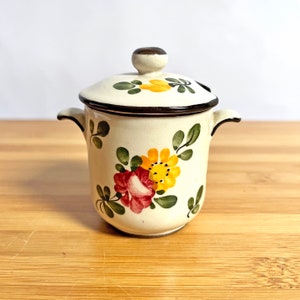 Peut inclure: Un petit pot en céramique blanche avec un couvercle, décoré d'un motif floral dans des tons de rouge, jaune et vert. Le pot a deux petites poignées sur les côtés.