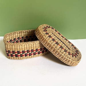 May include: An oval-shaped woven basket with a matching lid, crafted from natural tan fibers. The basket features a repeating geometric pattern in dark brown and red woven throughout the sides and the top of the lid, creating a textured look.