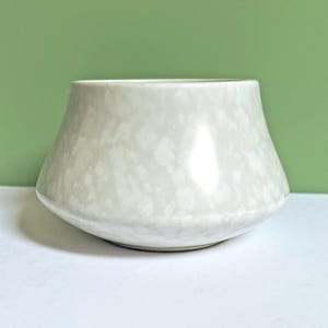 May include: A light-colored ceramic vase with a speckled pattern. The vase has a wide, rounded base that tapers upwards to a wider opening. The background is a solid green color.