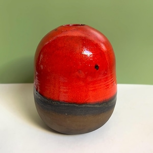May include: A small, round ceramic vase with a red glaze and a black band around the top. The vase is sitting on a white surface.