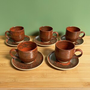 May include: Set of five red and brown ceramic coffee cups with matching saucers. The cups and saucers have a speckled glaze.