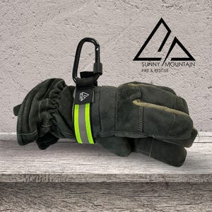 May include: Dark green work gloves with a black carabiner clip attached. The gloves feature a reflective yellow stripe and a black strap with a logo. The Sunny Mountain Fire & Rescue logo is visible in the background.