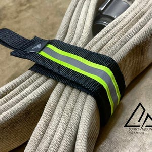 May include: Black and neon green reflective strap wrapped around a beige fire hose. The strap has a reflective silver stripe in the middle. The Sunny Mountain Fire & Rescue logo is in the bottom right corner.