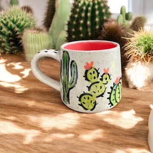May include: White ceramic mug with a red interior, featuring hand-painted green cactus designs and orange flowers. The mug has a speckled finish and a curved handle with black dots. The mug is set on a wooden surface, with cacti in the background.