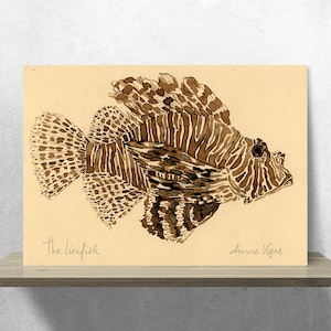 Op de afbeelding: Een bruine en witte aquarel illustratie van een koraalduivel met de tekst "The lionfish" en de handtekening van de kunstenaar "Anne Vigne" op een beige achtergrond.