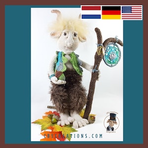 Puede incluir: Una figura amigurumi de crochet de una criatura del bosque con pelaje marrón, un chaleco verde y un bastón con un atrapasueños. La figura está sentada en una cama de hojas de otoño.