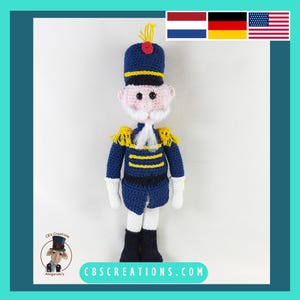 May include: A crocheted toy soldier wearing a blue uniform with yellow stripes, a white hat with a red pom-pom, and a white beard. The toy has a pink face with black eyes and a white mustache.