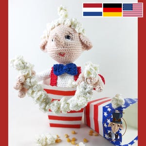 Könnte beinhalten: Eine gehäkelte Puppe, die ein rot-weiß gestreiftes Hemd, eine blaue Fliege und einen weißen Hut trägt. Die Puppe hält in jeder Hand eine Popcornkette. Die Puppe steht vor einer rot-weiß-blau gestreiften Popcornbox.