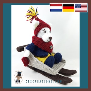 May include: A white crocheted teddy bear wearing a red and blue winter outfit sits in a gray crocheted sled with brown skis. The bear is wearing a red hat with a yellow and white pom-pom.