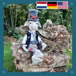 Könnte beinhalten: Ein grauer Häkel-Zwerg, der einen blauen Zylinder mit orangenen Schutzbrillen, eine schwarze Weste und einen grauen Schal trägt. Der Zwerg sitzt auf einem großen, verwitterten Baumstamm. Der Zwerg hält ein kleines, rot-weißes Objekt in der Hand. Der Hintergrund ist eine grüne Grasfläche mit Bäumen.